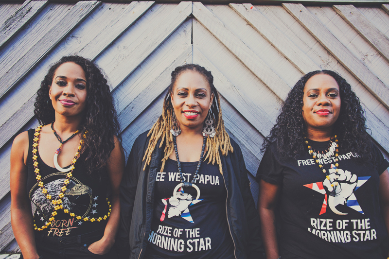 The Black Sistaz standing against wooden wall, smiling wearing Free West Papua t-shirts